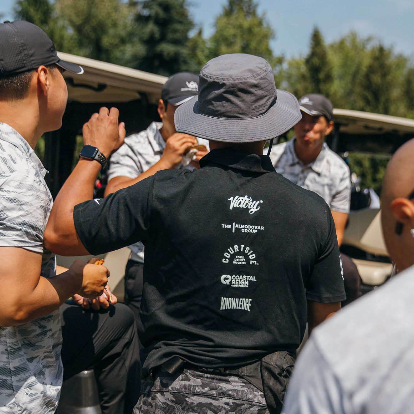 Black T-shirt featuring a vertically stacked custom printed design on the back with multiple white logos, including “Victory,” “The Almodovar Group,” “Courtside,” “Coastal Reign,” and “Knowledge,” shown being worn outdoors.