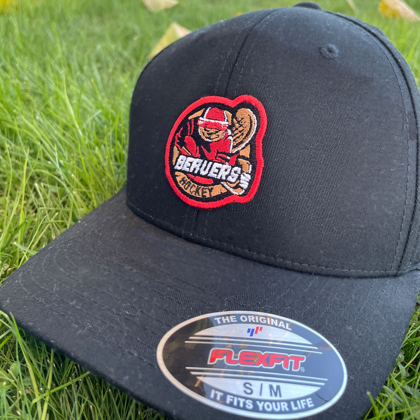 A close-up photo of a black Flexfit baseball cap resting on grass. The cap features an embroidered patch of a cartoon beaver in red hockey gear holding a hockey stick, with the words “BEAVERS HOCKEY” in white text outlined in red. A shiny oval Flexfit sticker on the brim reads, “THE ORIGINAL FLEXFIT S/M IT FITS YOUR LIFE.”