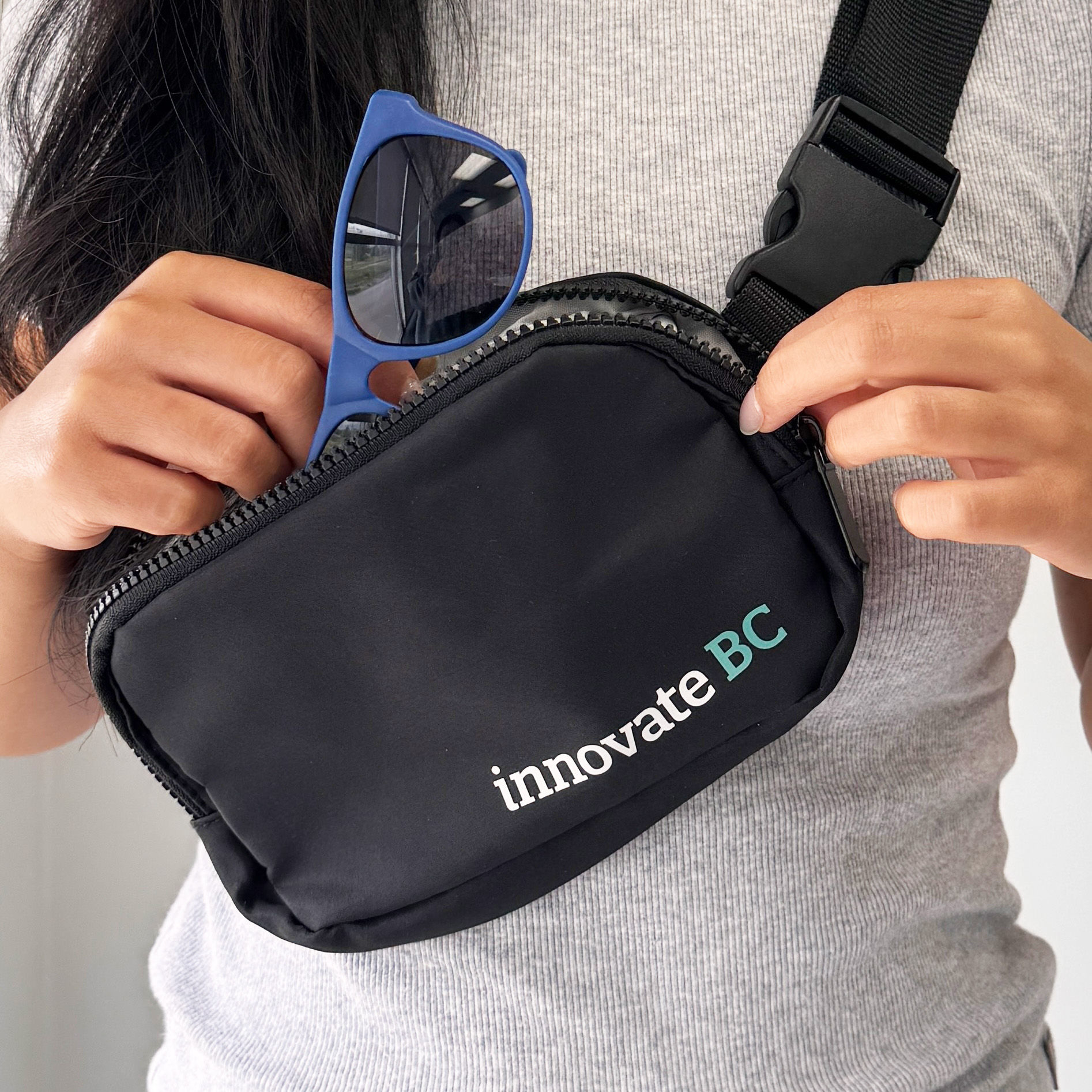Branded Innovate BC black fanny pack worn crossbody, with person placing blue sunglasses inside.