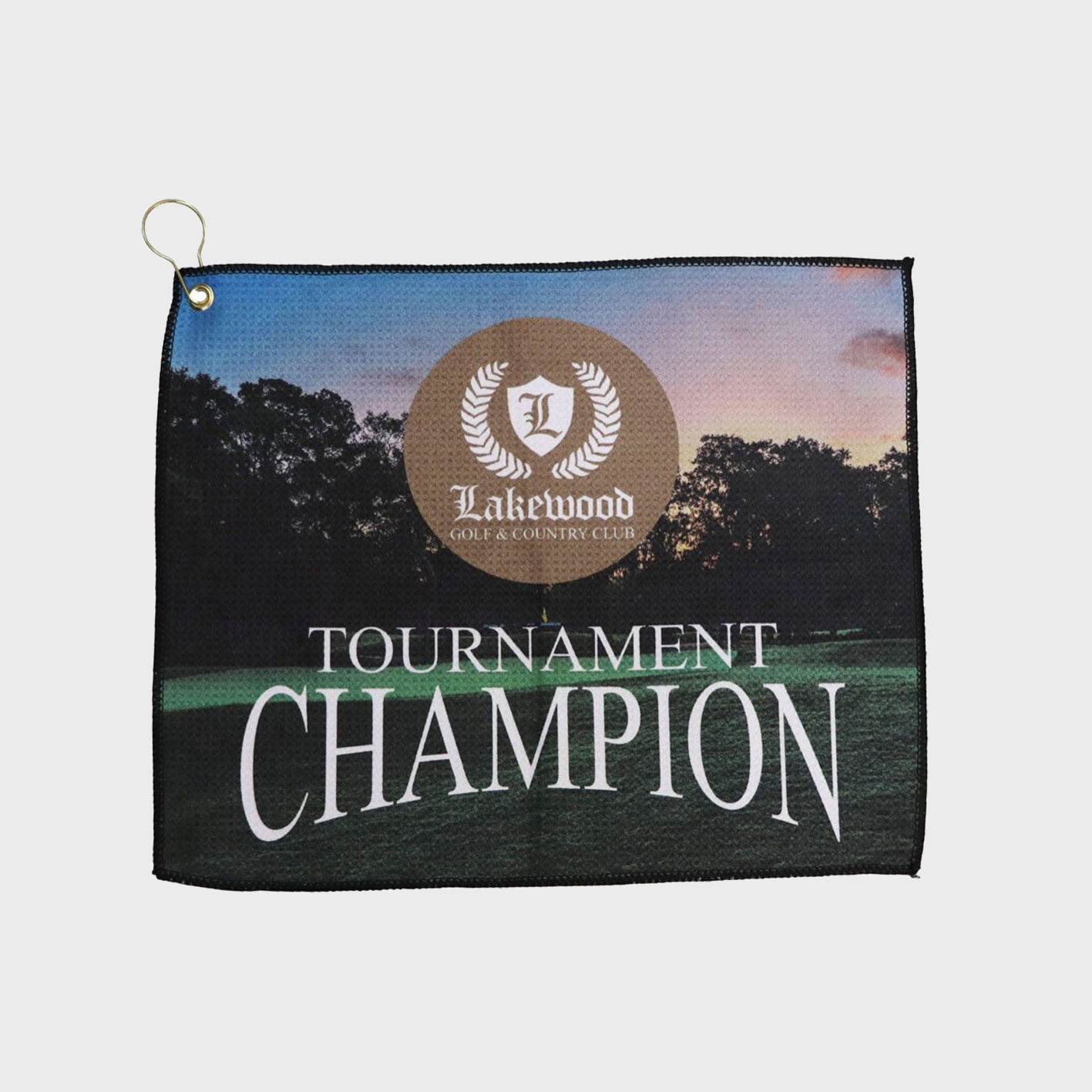 Custom printed golf towel with Lakewood Golf & Country Club logo and the words ‘Tournament Champion,’ featuring a sunset golf course background and a grommet with hook in the corner.