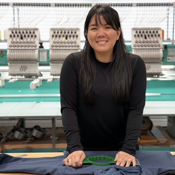 A smiling young woman in a black long-sleeve shirt stands in front of a multi-head embroidery machine, with a bright, clean background of white window blinds and light teal work tables. She is holding a green plastic centering tool over a folded navy-blue sweatshirt on a wooden table in the foreground, ready to start the embroidery process.