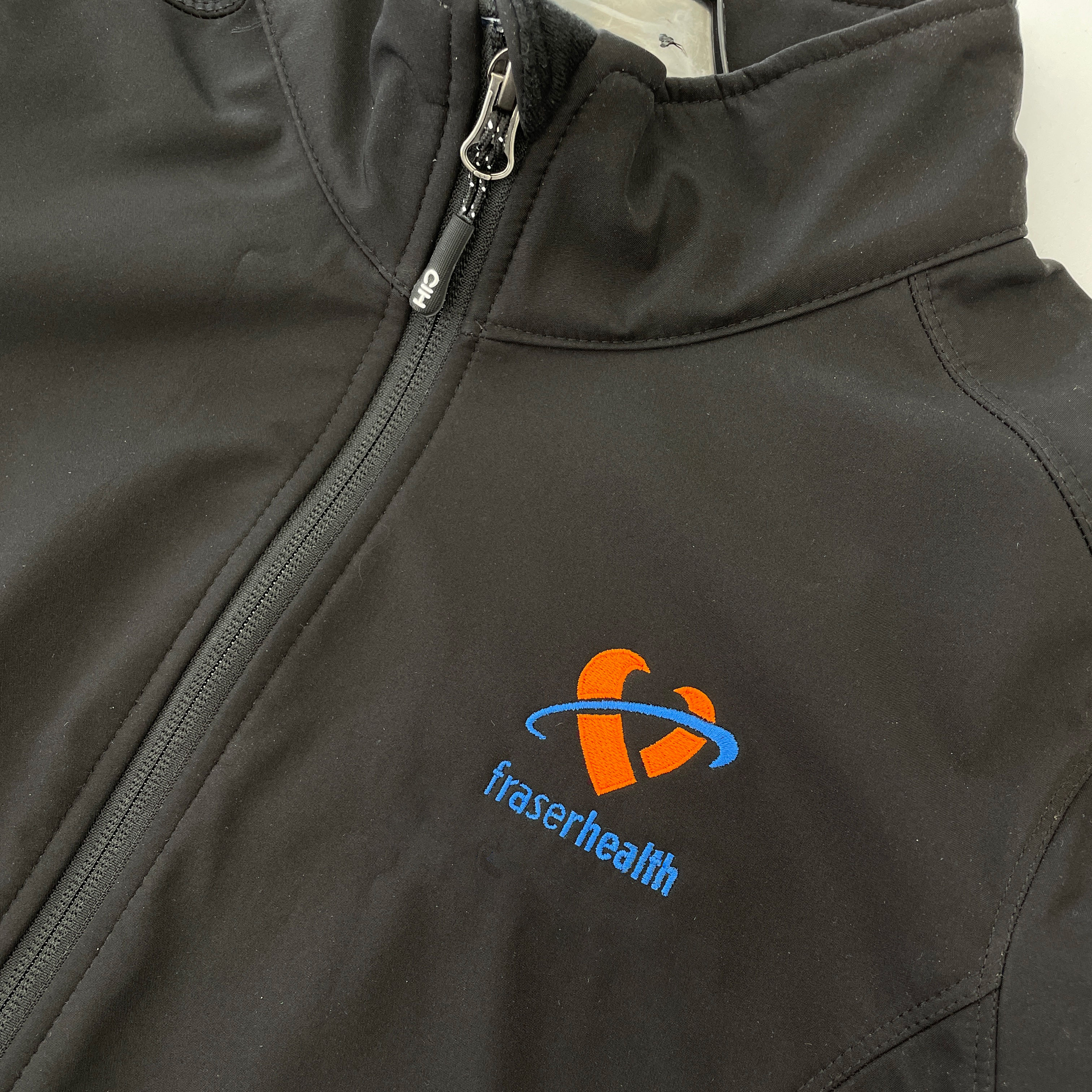 Fraser Health branding stitched in white and orange thread on the chest of a custom black softshell jacket.