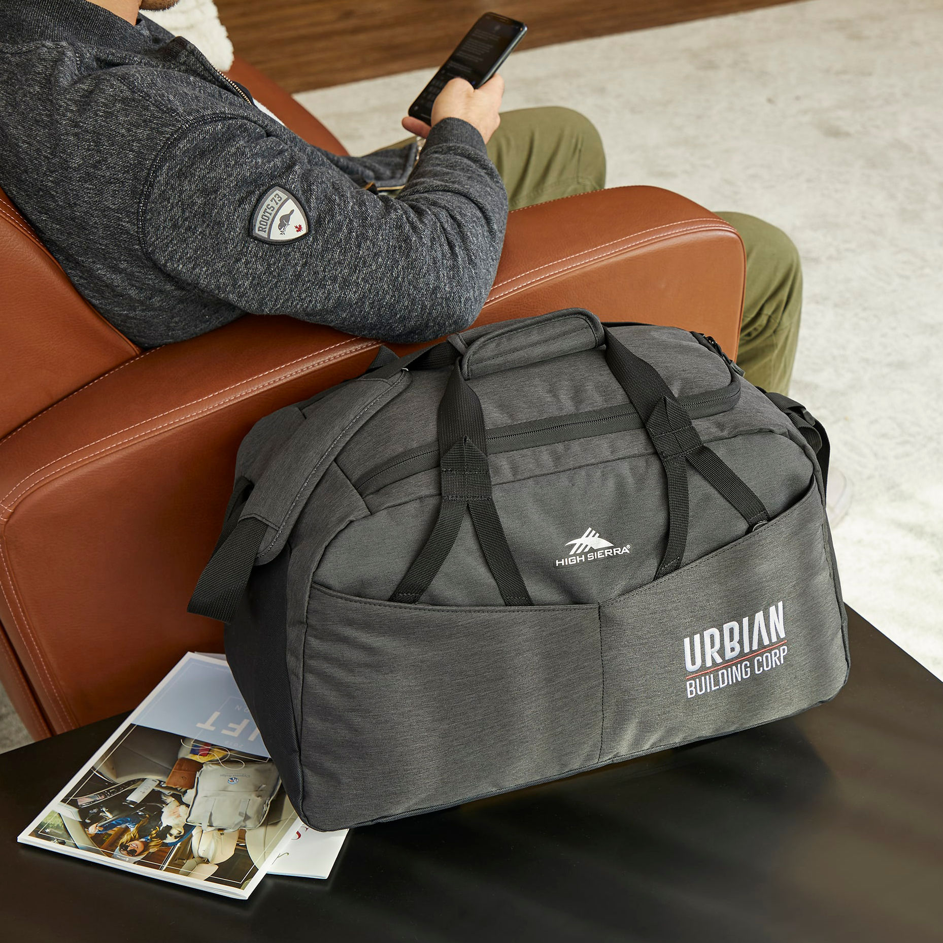 Gray duffel bag with a custom printed “URBIAN Building Corp” logo in white on the front pocket, shown in a modern lounge setting with a High Sierra brand mark above the handles.