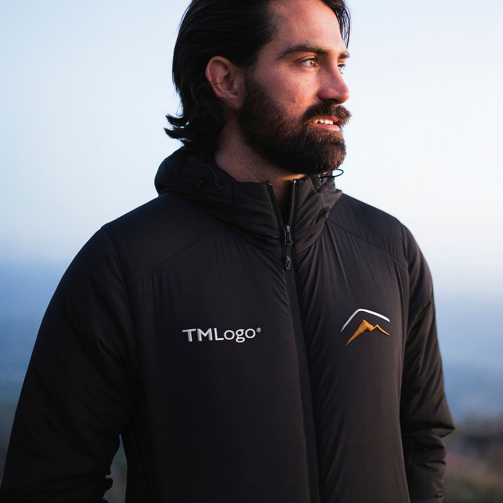 A man with a beard and long dark hair stands outdoors. He is wearing a black jacket featuring two embroidered-style logos on the chest—one on the left with the text “TMLogo®” and one on the right depicting a stylized mountain design in gold and white.