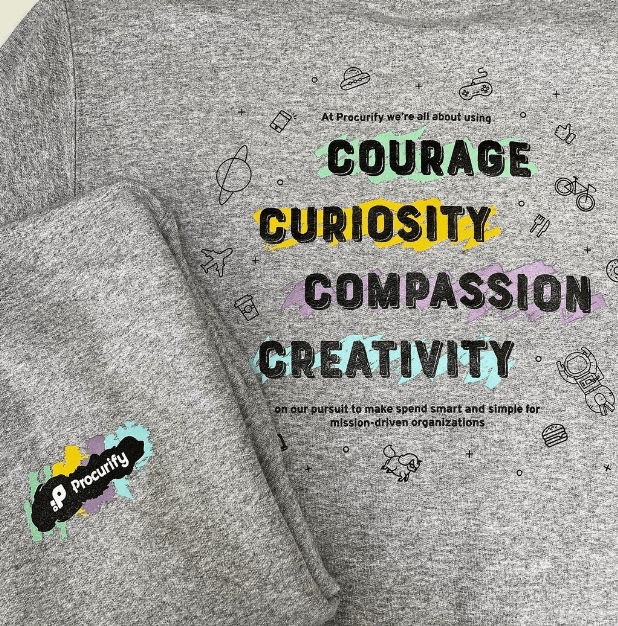 Gray heather T-shirt featuring a colorful custom printed typographic design with the words “Courage,” “Curiosity,” “Compassion,” and “Creativity,” surrounded by small illustrated icons, with a Procurify logo printed on the sleeve.
