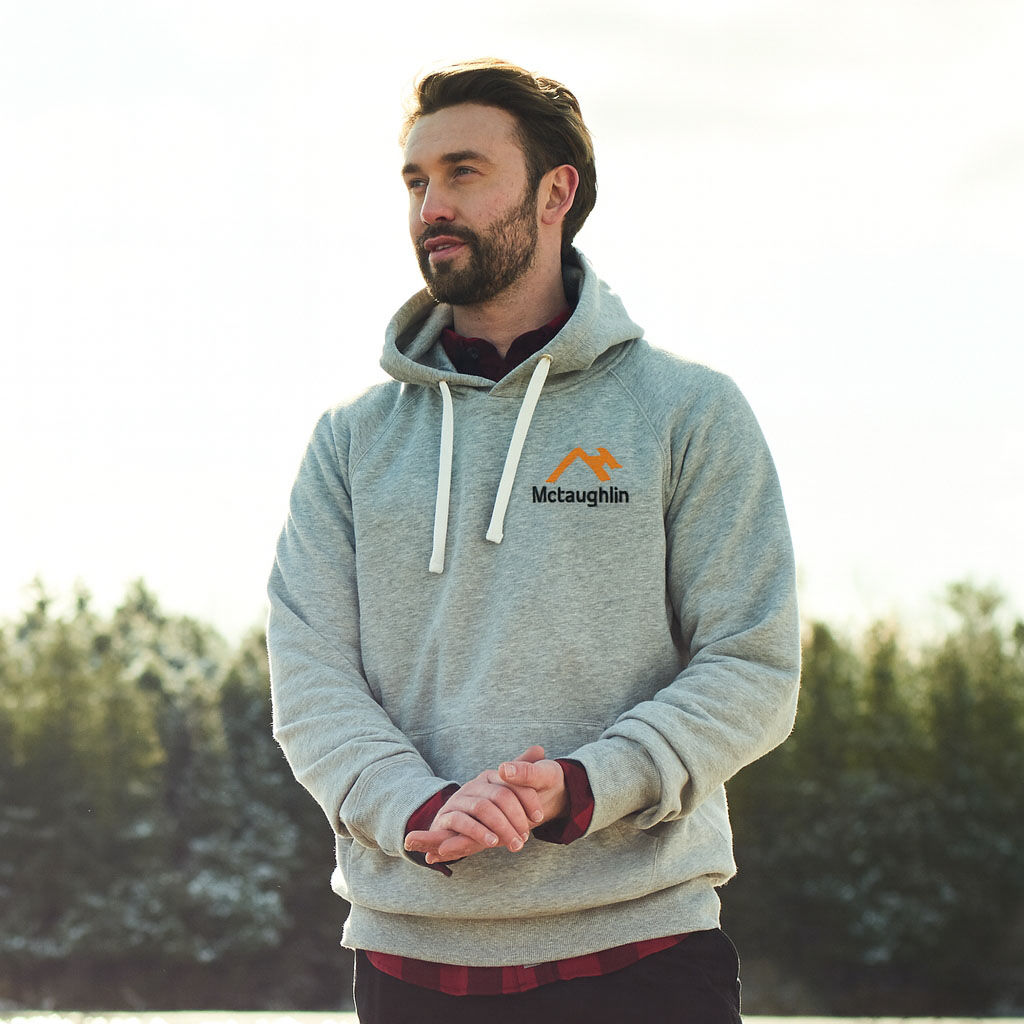 A man wearing a light grey McLaughlin Construction hoodie with white drawstrings and an embroidered company logo on the left chest, standing outdoors in front of snow-covered evergreen trees.